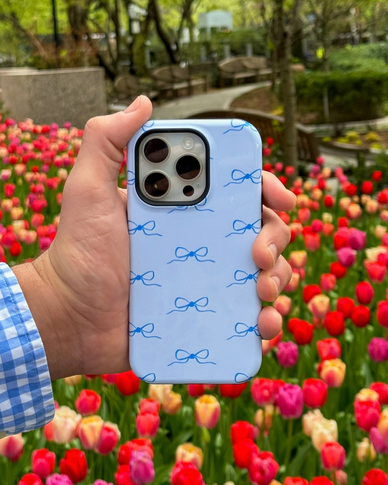 Hand holding a phone with a blue case featuring dark blue bows against a colorful tulip garden background